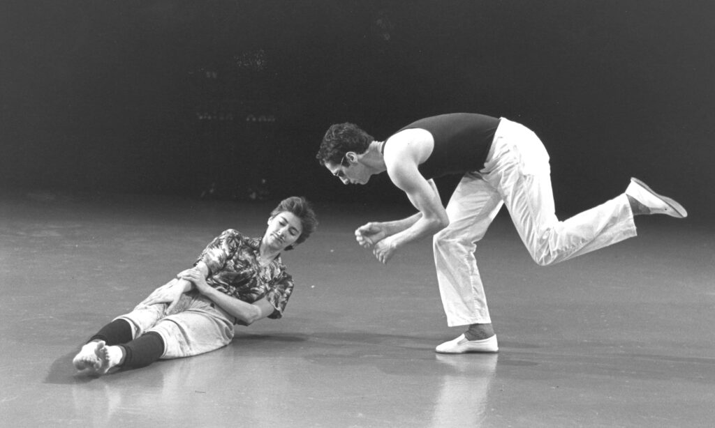 In a black and white archival image, Steve Paxton faces the side as he balances on one leg, torso parallel to the ground as his free leg bends behind him as though running. Lisa Nelson lies on the floor a couple of feet in front of him, lifting the top of her torso slightly off the ground to slide her right hand down her thigh.