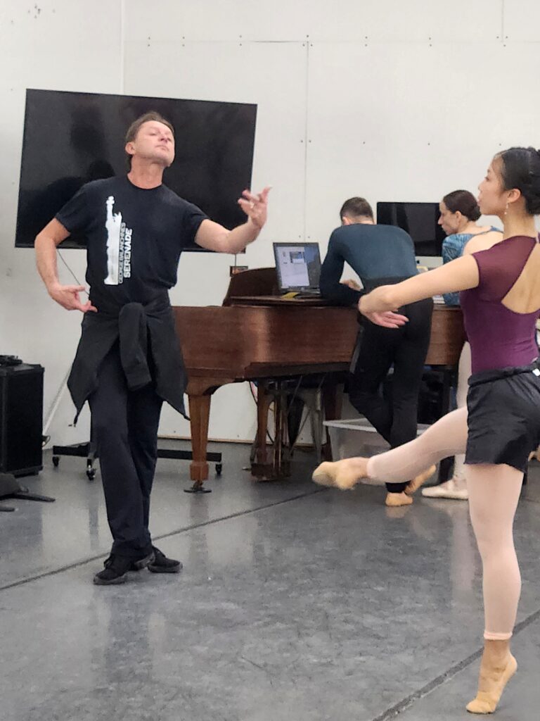 In a dance studio, Timour Bourtasenkov gestures with his left hand toward dancer Katharine Cowan.
