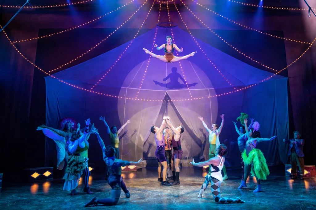 A dancer flies high above the stage in a toe touch as a trio stands below waiting to catch her. Eight elaborately costumed circus performers form a circle around them, all facing in and up.