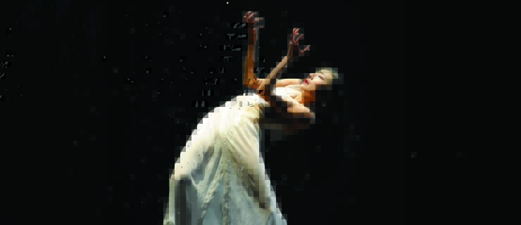 Yuan Yuan Tan stands flat footed, arching back so her spine is nearly level with the ground as she stares at her upraised hands, ash falling onto and around her. Her dark hair is loose and falls behind her; she wears a white dress that drapes to the dark stage and matching white pointe shoes.