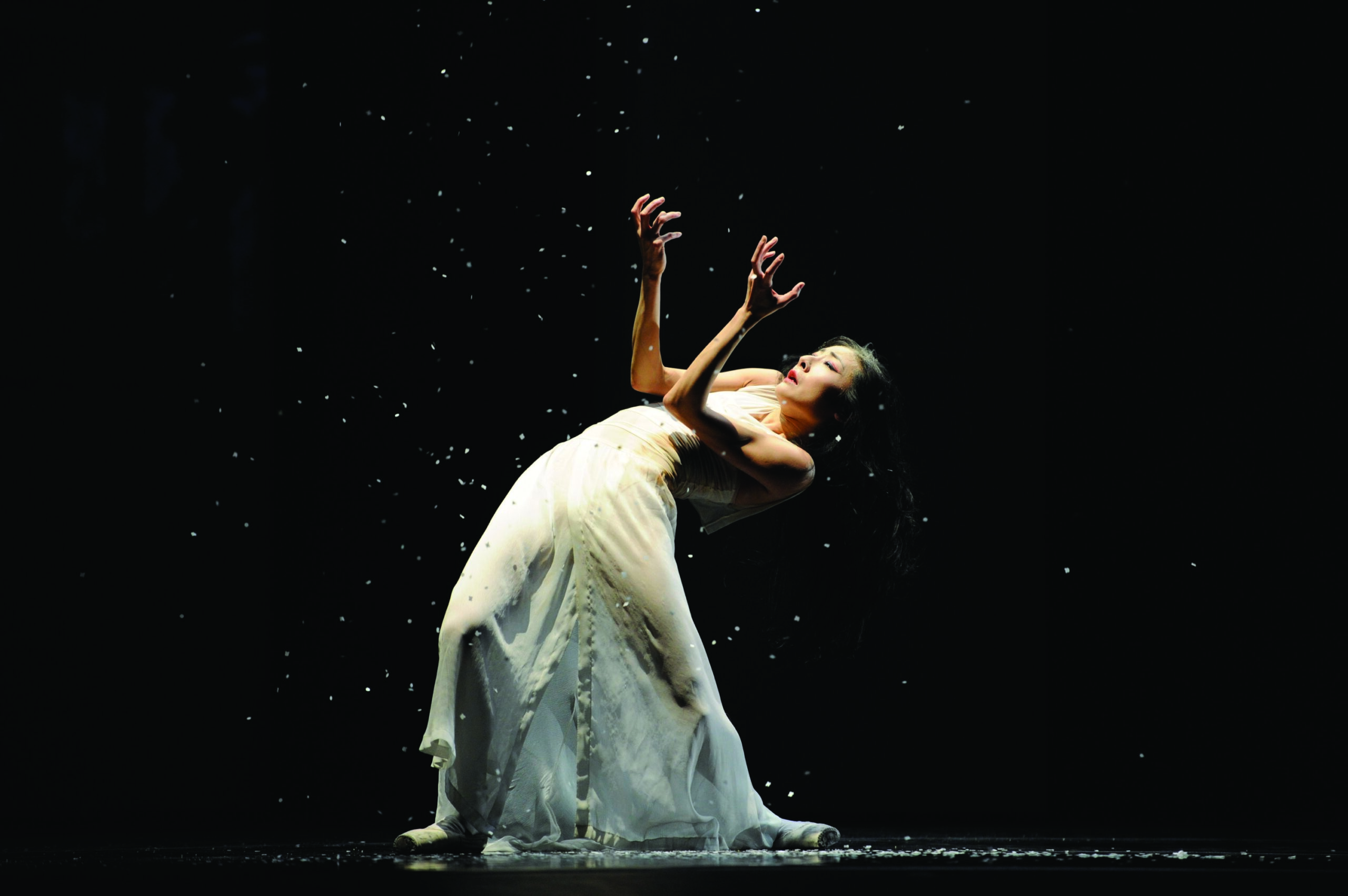 Yuan Yuan Tan stands flat footed, arching back so her spine is nearly level with the ground as she stares at her upraised hands, ash falling onto and around her. Her dark hair is loose and falls behind her; she wears a white dress that drapes to the dark stage and matching white pointe shoes.