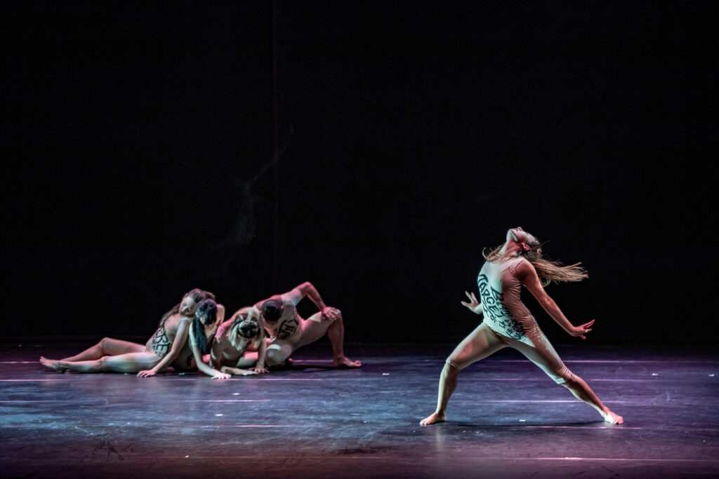 a group of dancers huddled together update with one female dancer standing downstage