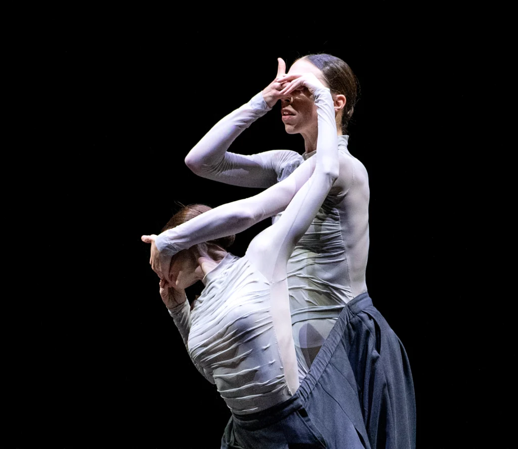 Two female dancers on a darkened stage stand close to each other, sharing the same pair of pants. Their arms intertwine as their hands meet and cover each other's eyes. The downstage dancer is in plié, twisting upstage toward the dancer standing tall just behind her.