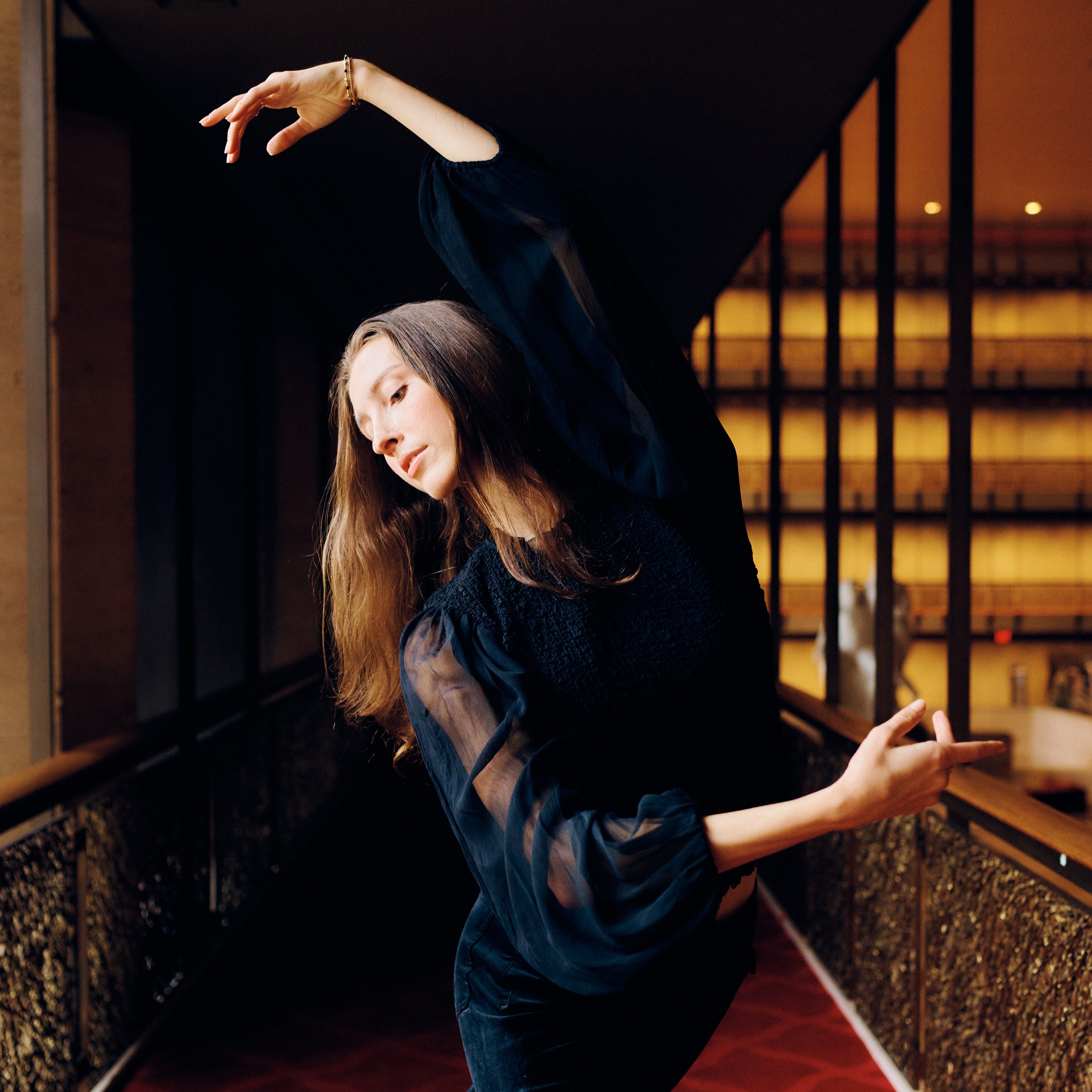 a female dancer wearing a blue long sleeved shirt stretching to the side with her arms in 4th position