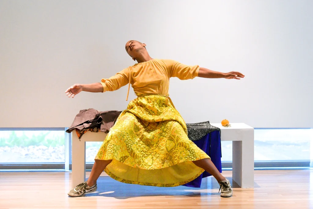 Artist Jasmine Hearn sitting on a white bench in front of a white wall in a gallery setting. They are wearing a brown blouse and a yellow skirt and tennis shoes. They are leaning back with both arms up and outstretched.