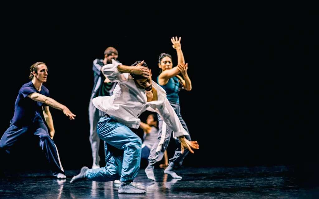On a dark stage, a dancer slides toward the floor, one hand blurred as it reaches for the ground and the other pulling his head to one side. Four dancers similarly costumed in sweatpants and different shirts are blurs of motion upstage.