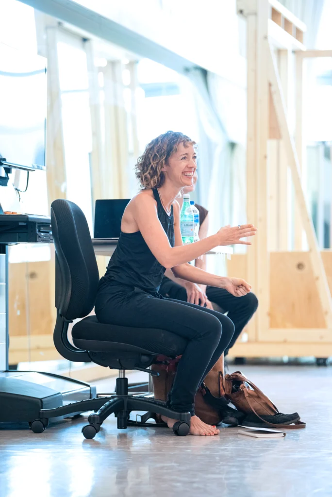 Cathy Marston smiles widely as she sits in a rolling chair at the front of a sunny, mirrored rehearsal studio. She is barefoot, a notebook sitting at her feet.
