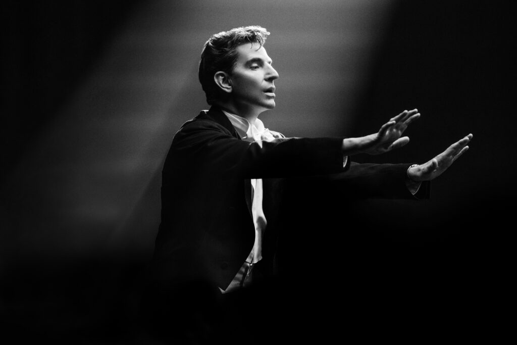 A black and white photo of Cooper as Bernstein at the podium, wearing a tuxedo. His arms are raised, his expression intent.