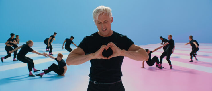 Gosling, wearing a black tee and jeans, makes a heart with his hands in front of his chest. An ensemble of identically-dressed men dance behind him.