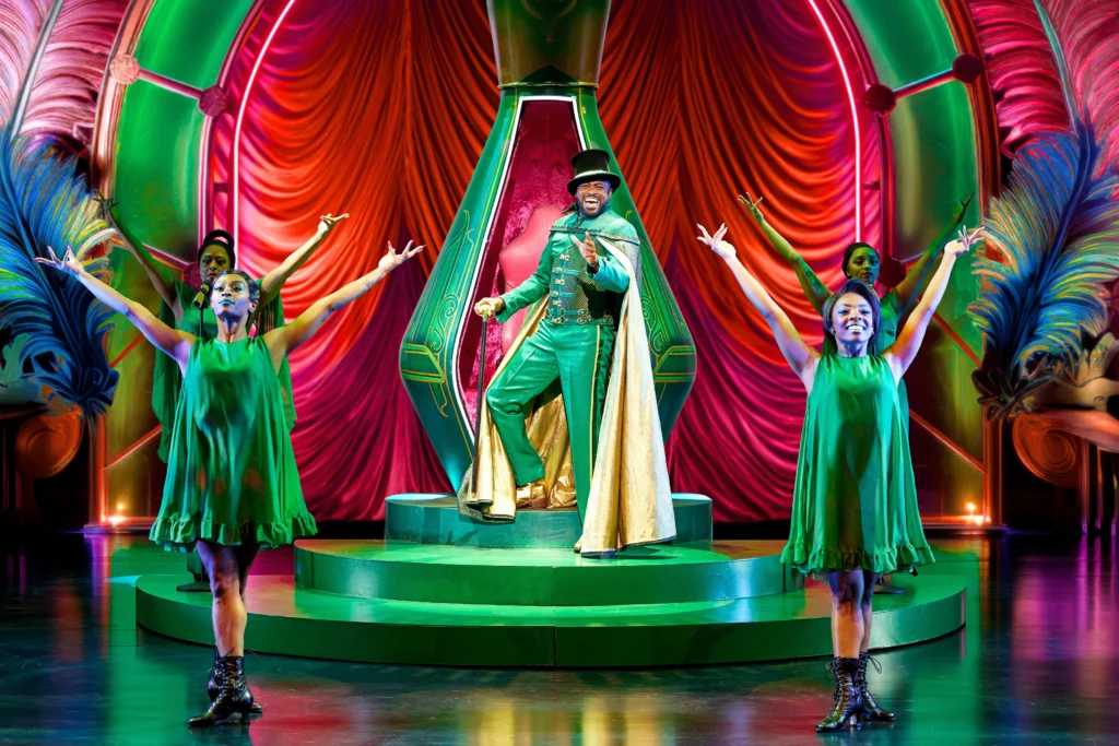 A green and gold garbed Wayne Brady as The Wiz. He stands before a red and green throne, singing out to the audience. Four dancers face out to the audience, palms out and up.