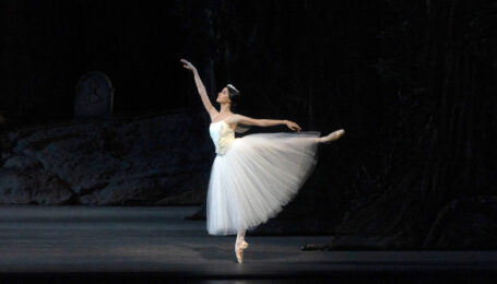 a female dancer wearing a long white tutu performing in first arabesque on stage