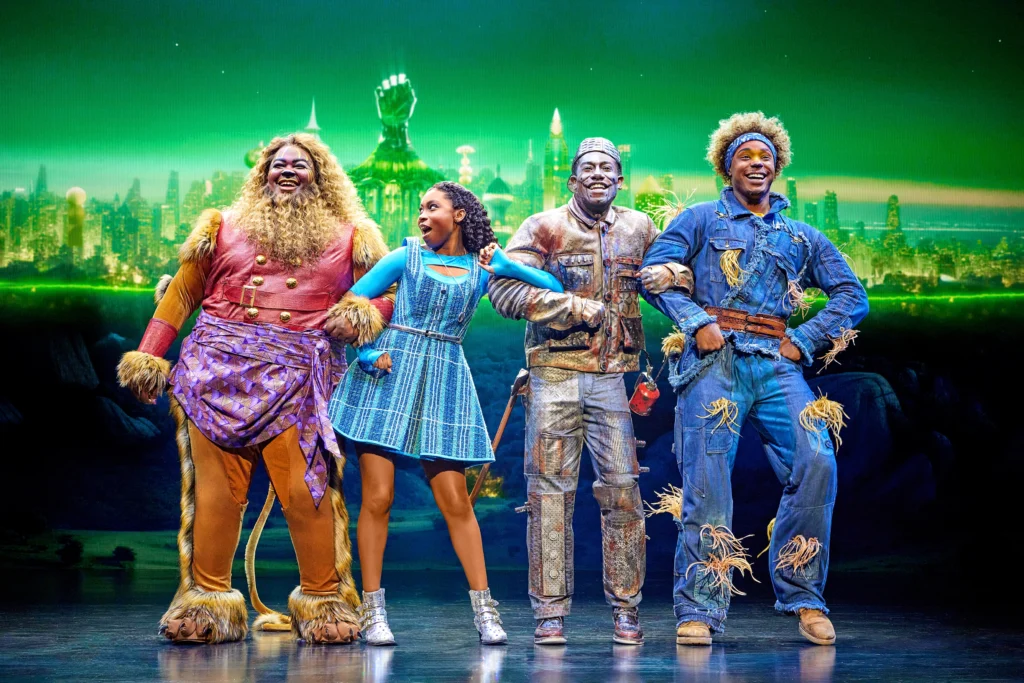 Four dancers in costume as the Lion, Dorothy, the Tin Man, and Scarecrow stand side-by-side in a line, arms linked in classic Wizard of Oz fashion. The Emerald City is visible in the background.