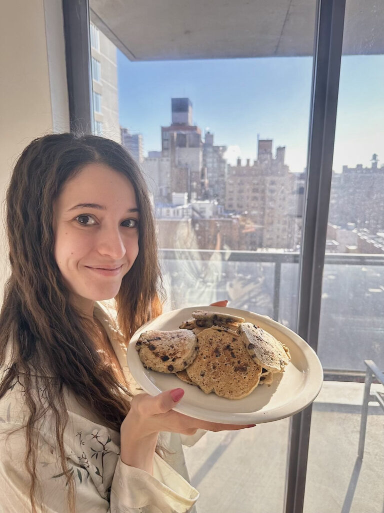 a woman holding a plate of pancakes standing next to a window with a skyline in the background