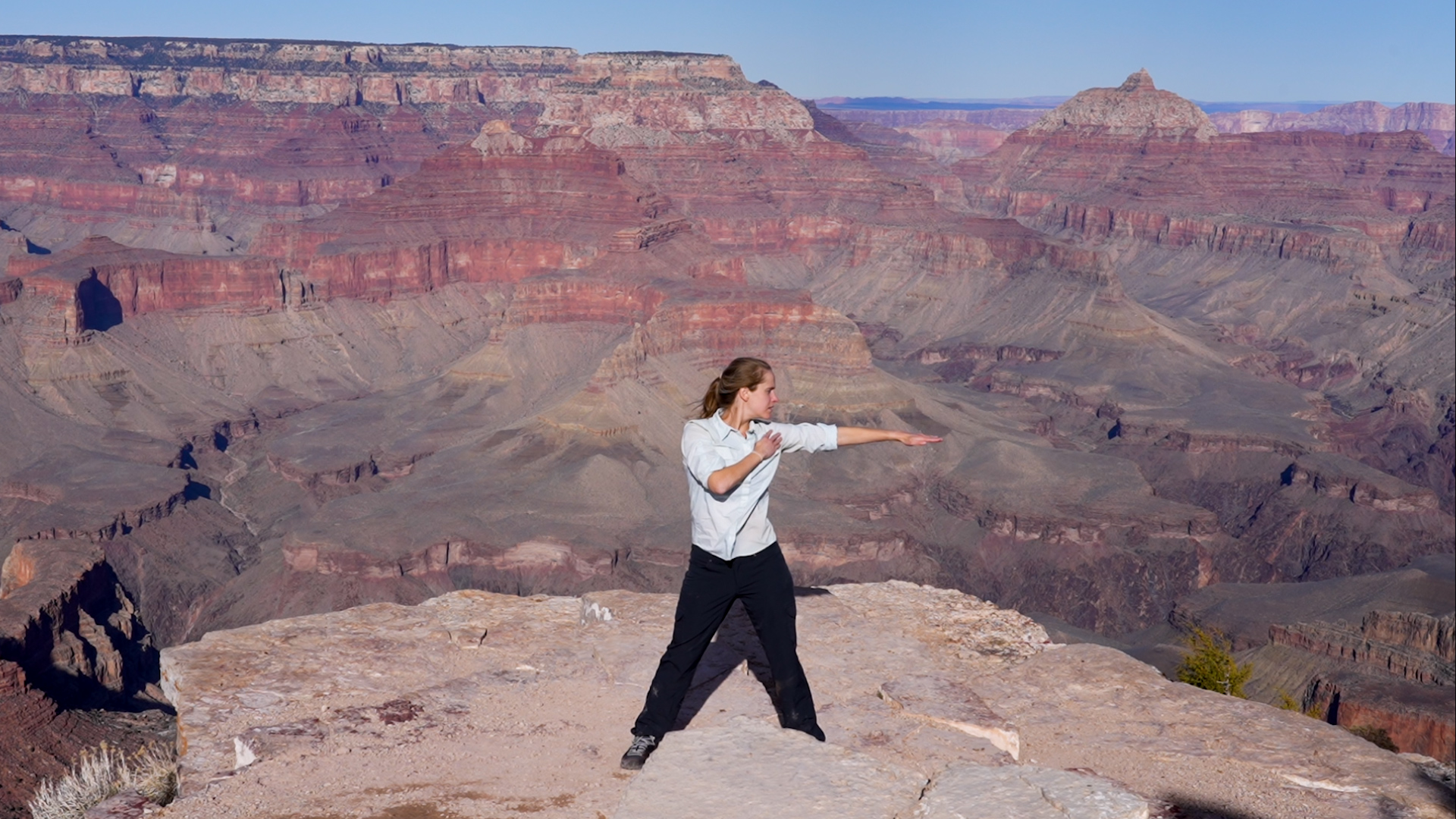 Denton, wearing a loose white shirt and black pants, stands at the rim of the Canyon on a brilliantly sunny day, her legs planted in a wide second position, looking out over her extended left arm.