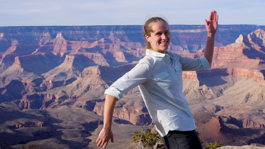 Denton, wearing a loose white shirt and black pants, stands at the rim of the Canyon on a brilliantly sunny day, smiling into the camera, her elbows forming right angles, with her left hand pointing to the sky and her right to the ground.