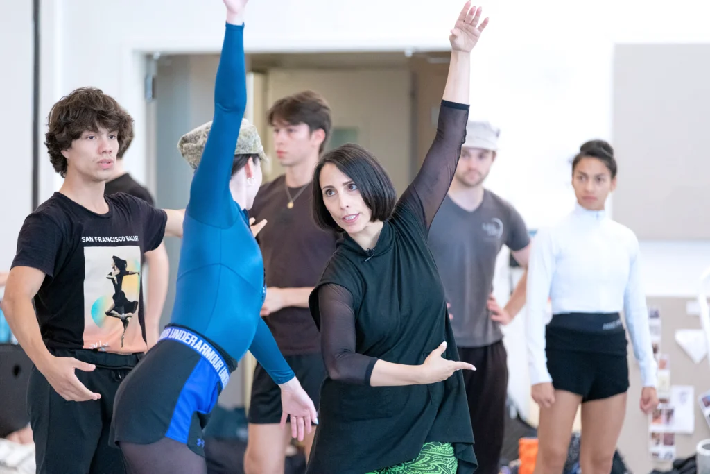 Annabelle Lopez Ochoa demonstrates a pose, one arm raised as the other wraps toward her waist, as a dancer mirrors her, others crowding around watching.