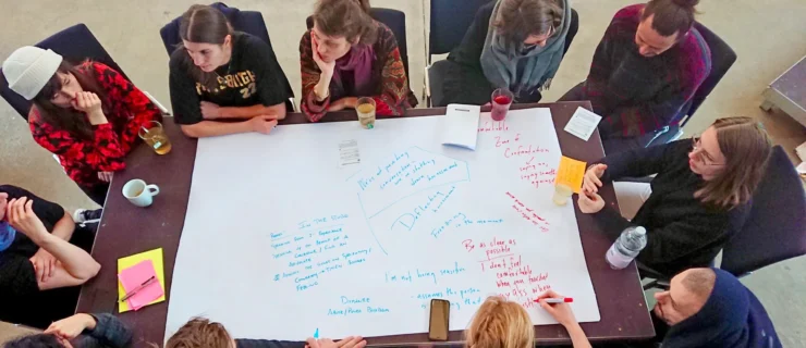 Roughly a dozen people sit crowded around a table that is mostly covered in a large sheet of paper strewn with red and blue writing. Someone adds to it in red marker while they listen to each other speak.
