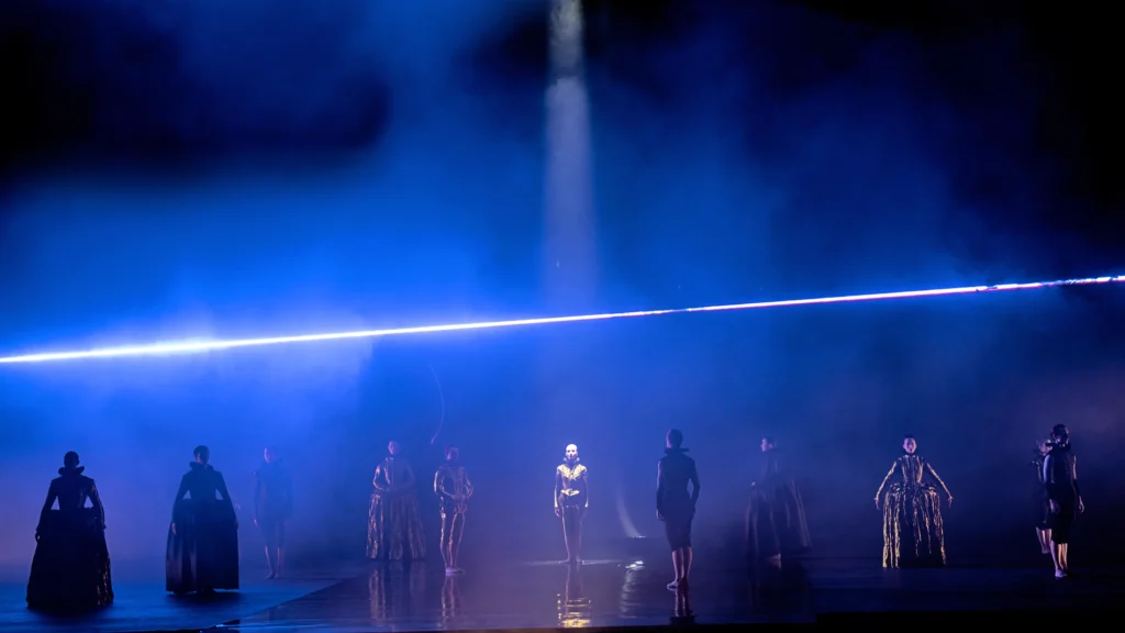 A massive, foggy stage is lit blue as a laser of light cuts the space from stage left to stage right. Ten dancers are scattered around, facing different directions, wearing neck ruffles and, in some cases, broad skirts. A singular dancer is spotlit, upstage center, facing downstage.