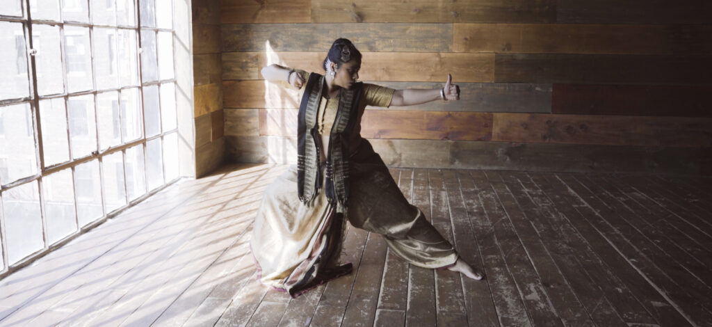 a female dancer dressed in traditional clothing moving in an open room with wood paneling and large windows 