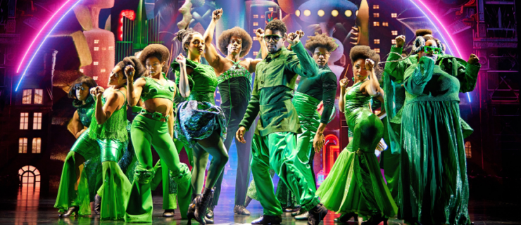 Nine dancers in green cluster together and pose mid-snap beneath a technicolor skyline.