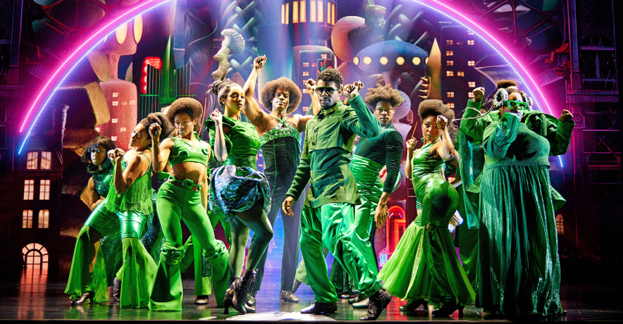 Nine dancers in green cluster together and pose mid-snap beneath a technicolor skyline.