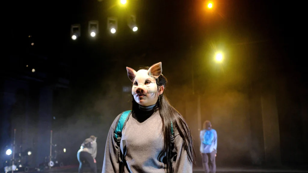 A dancer stands downstage, shown from the waist up, the top half of their face hidden by a pig mask. Their hair is straight black and loose to their elbows. They wear a backpack. Two dancers are blurry upstage.