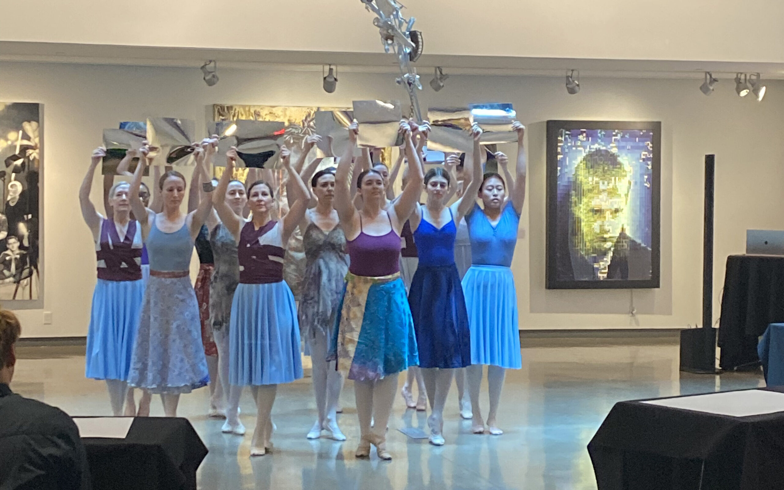 a group of adult female dancers huddled together holding shiny square sheets above their heads while dancing in an art gallery 