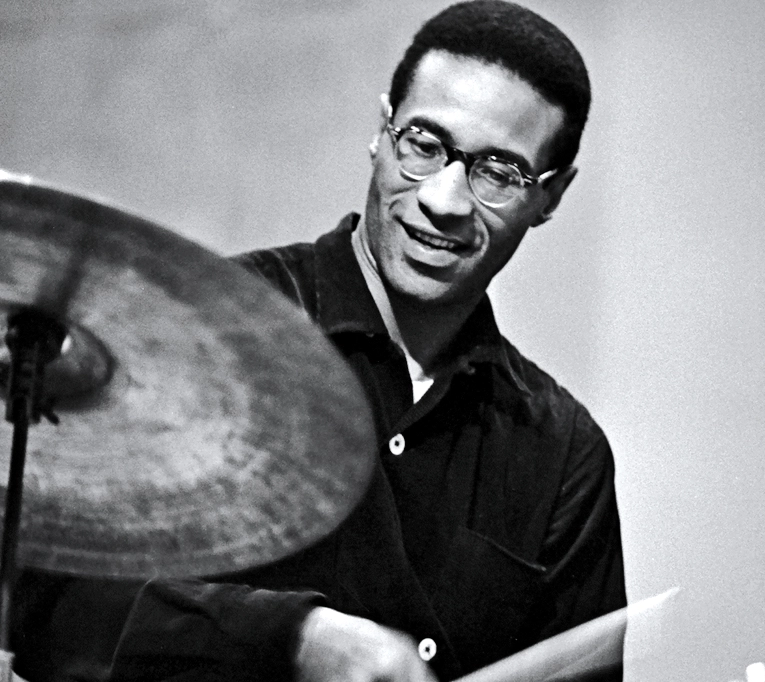 A black and white archival photo of Max Roach, smiling as he sits at a drumkit.