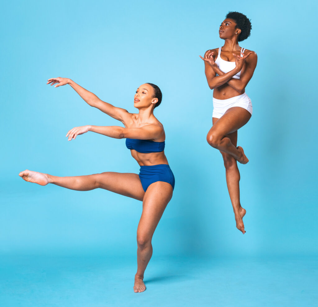 Two dancers pose against a teal backdrop. One extends her upstage leg to 90 degrees, arms in an extended third position. The other is caught midair, one foot tucked behind the opposite knee, arms crossed over her chest as she looks over one shoulder. Both are barefoot and wearing matching trunks and bra tops.