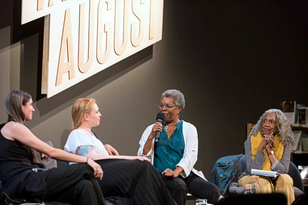 Four women sit onstage, holding microphones. Robyn Doty and Frances Chiarini, two young white women, are seen in profile as they turn toward Elisabeth Clarke-Hasters, an older Black woman, as she speaks. Brenda Dixon-Gottschild leans forward as she listens intently on Clarke-Hasters' other side.