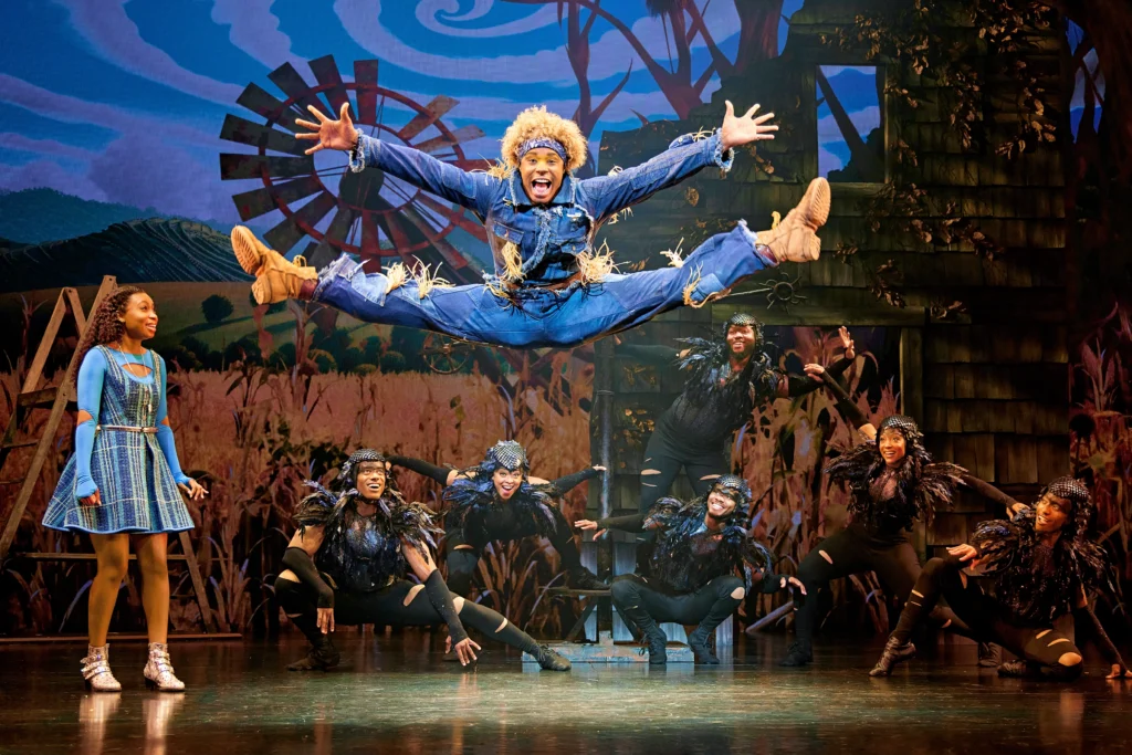 Avery Wilson is caught midair in a long, enthusiastic toe-touch. His arms are outstretched, palms open to the audience. He wears head to toe denim, beige boots, and a headband beneath fluffy yellow-orange hair. A half-dozen black-garbed dancers crouch upstage and look up at him with expressions of delight.