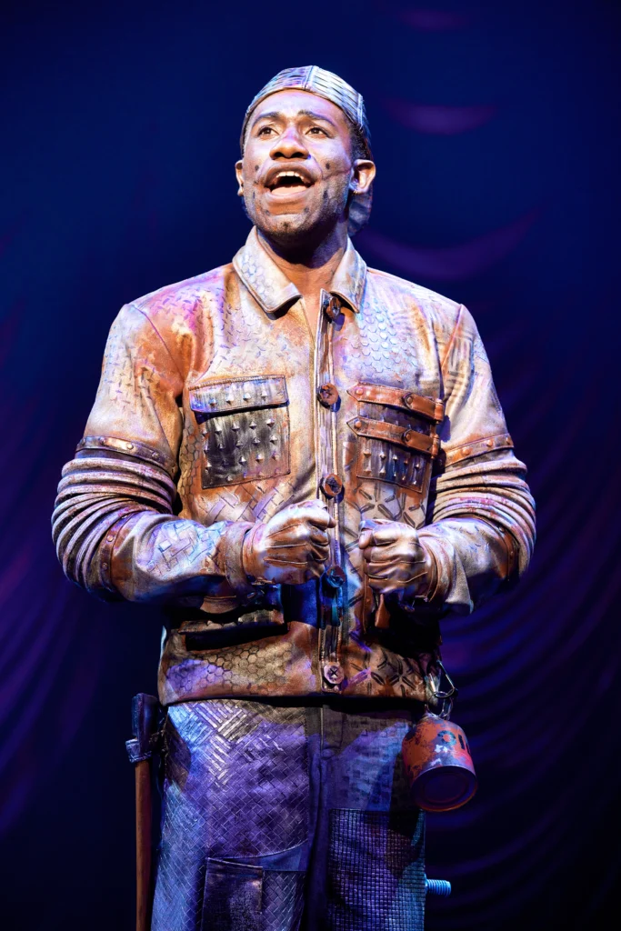 Phillip Johnson Richardson stands and sings as the Tin Man in The Wiz. He is painted silver, though his brown skin shines through, and wears a silver-painted backwards baseball cap and workman's jacket.