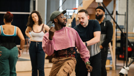 Kelly, wearing a pink cropped sweatshirt and olive baseball cap, demonstrates a step at the front of a studio full of dancers.