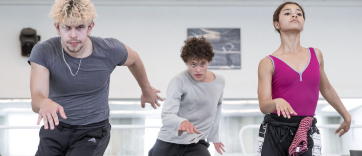 Three dancers in rehearsal-wear move through their hips in a way evocative of salsa dancing as they face forward.