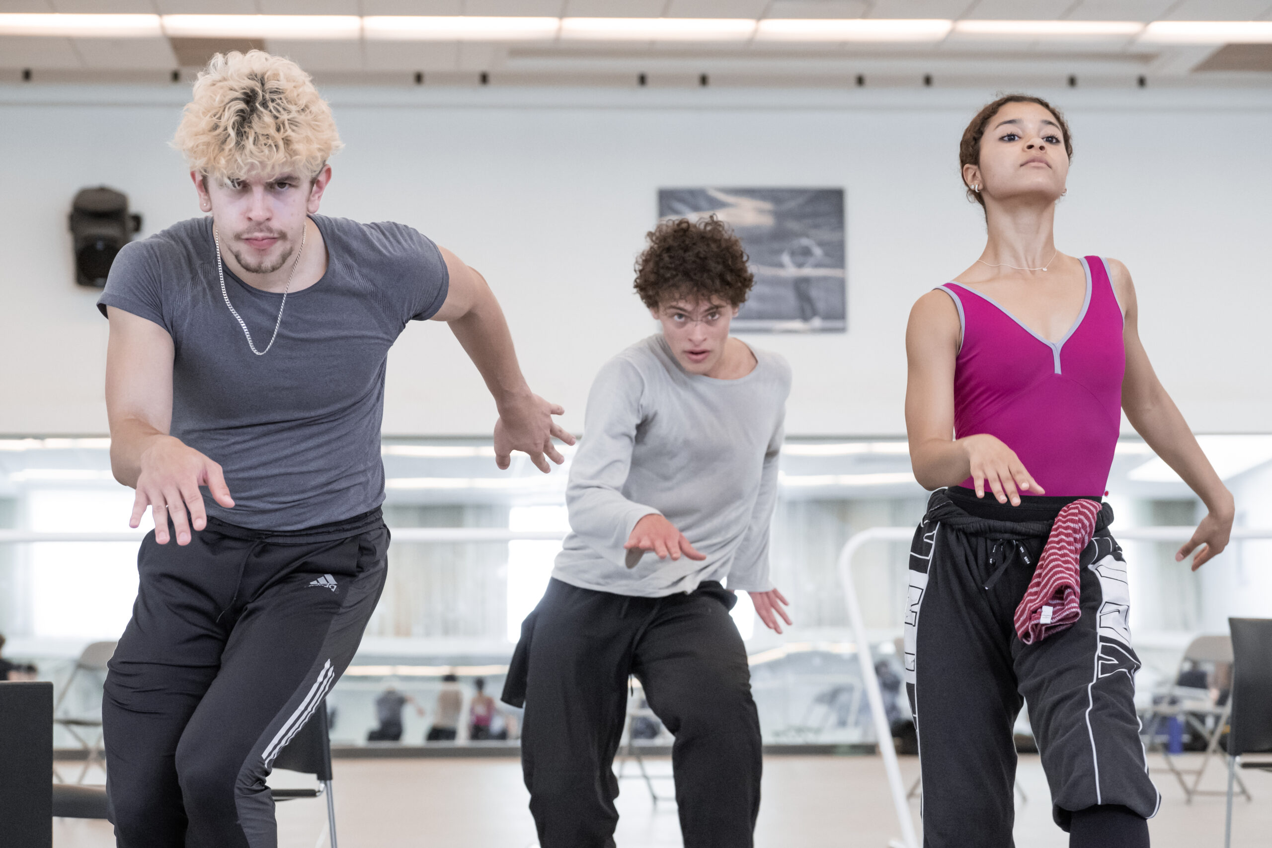 Three dancers in rehearsal-wear move through their hips in a way evocative of salsa dancing as they face forward.