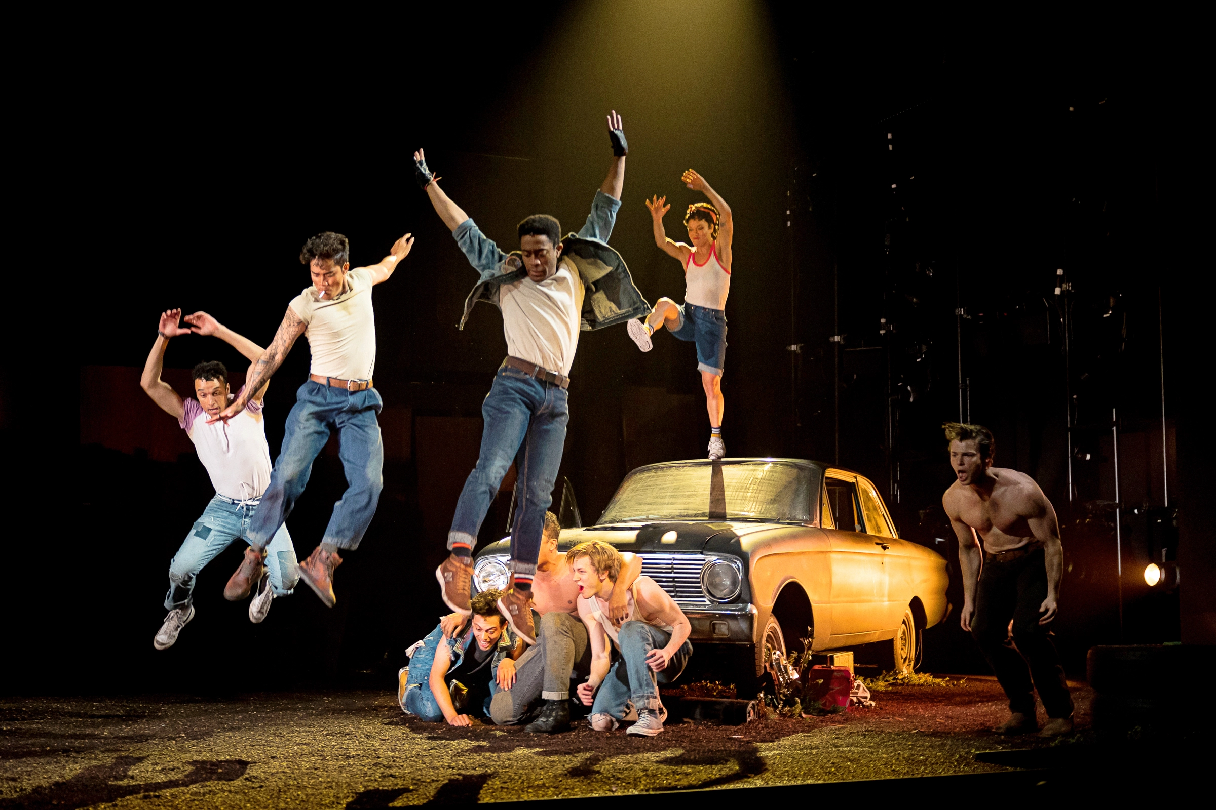 Eight performers move around a parked, old-fashioned car. One leaps from the top of the hood; a trio crouches together in front of the car. Another three