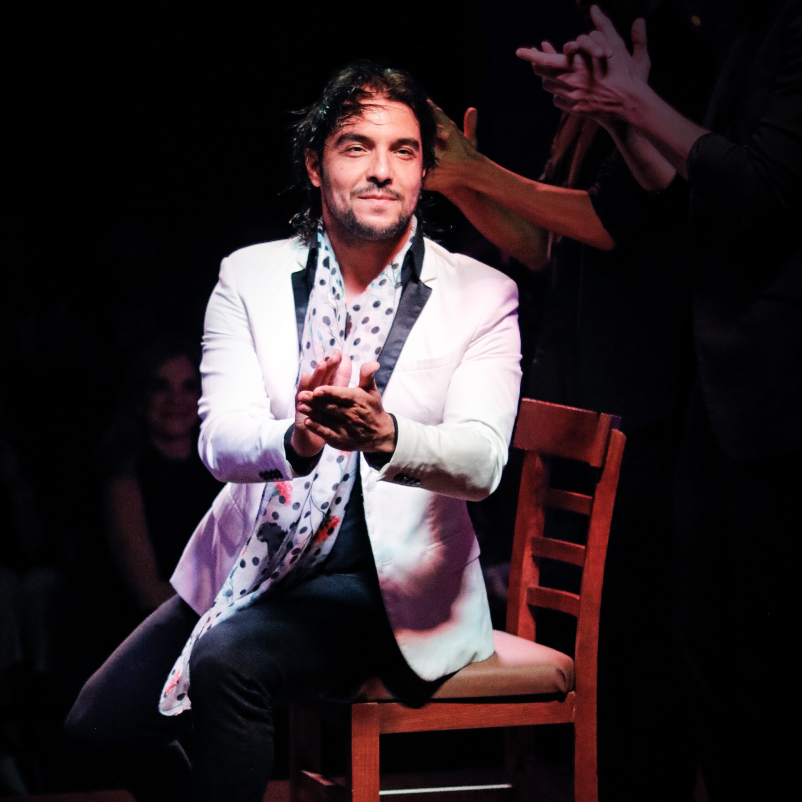 A man wearing a white jacket and polka dot scarf sitting in a chair clapping