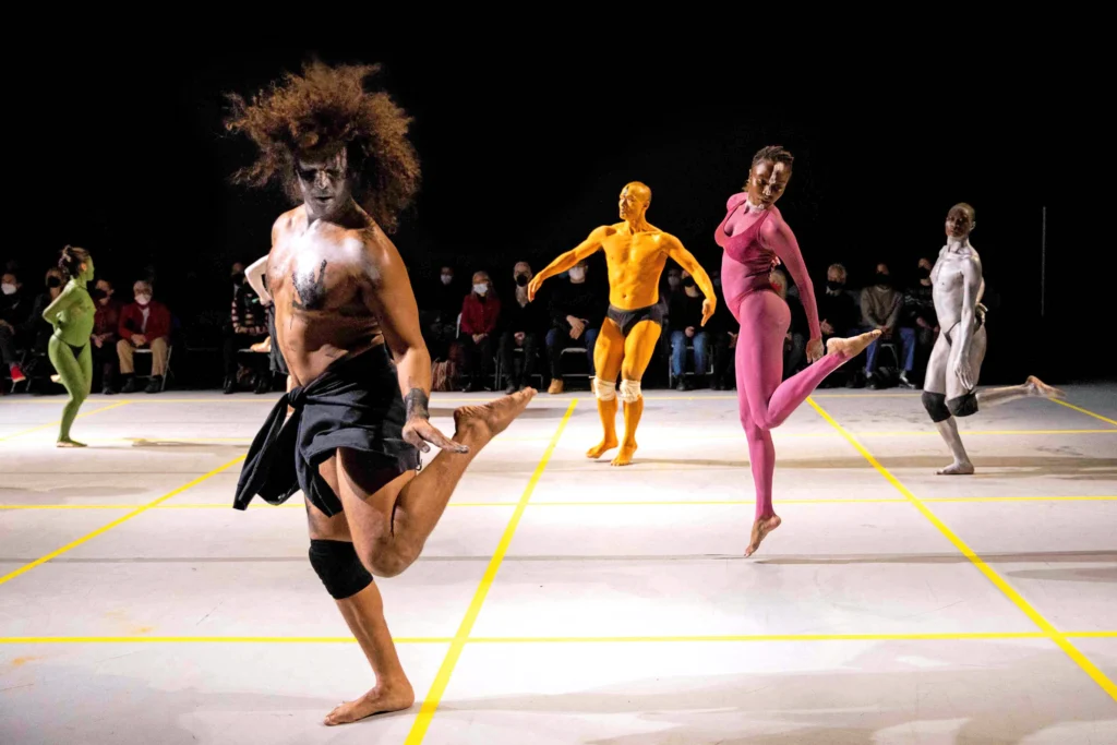 Five dancers painted bright colors dance spaced far apart, each holding to a square created by yellow tape on a white floor.