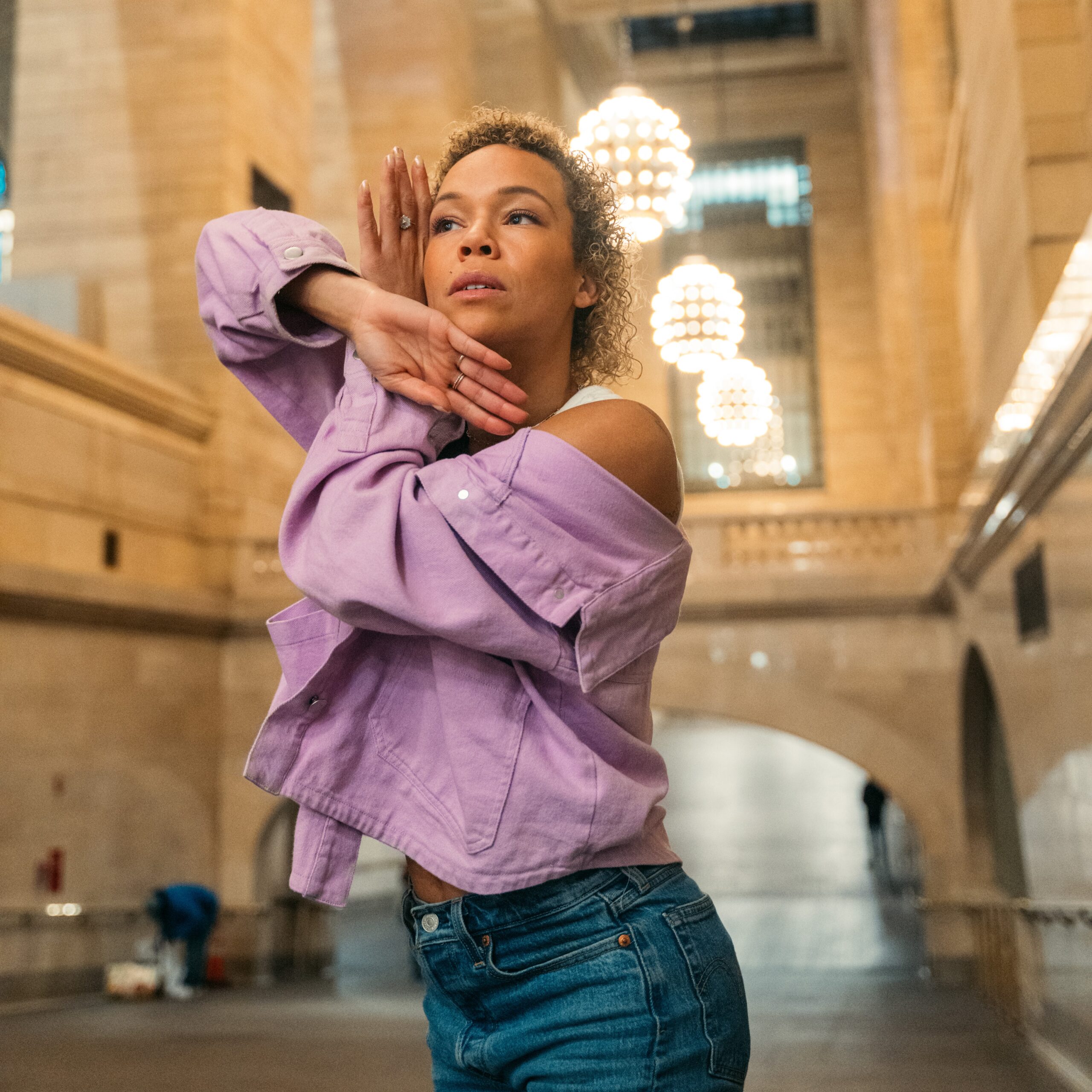 a female dancer wearing a purple button down shirt and jeans dancing in a large open room. she poses with her arms crossed and her hands near her face