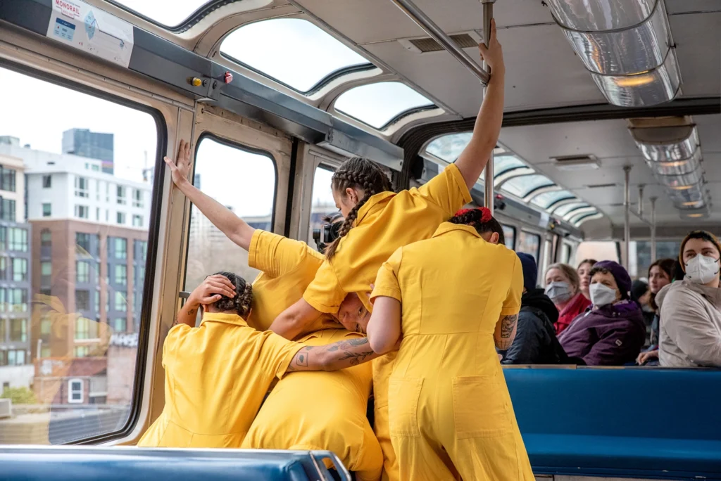 Four dancers in bright yellow cluster and connect with their backs to the camera as they perform on a public train.