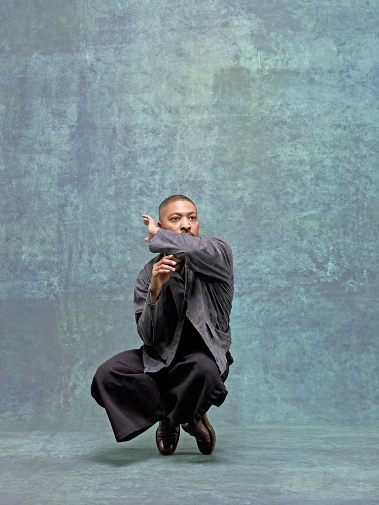 Kyle Abraham poses against a blue backdrop. He crouches, balancing on the toes of his shoes, arms wrapping around his head.
