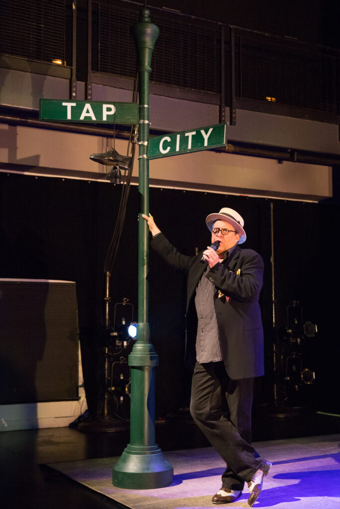 Tony Waag speaks into a handheld microphone while leaning against a green signpost, which holds signs reading "Tap City."