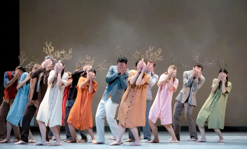 A dozen dancers in different colored dresses and shirt and pants combinations hide their faces behind their hands, leaning as one to the right. Branches sprout from their heads.