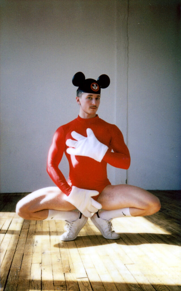Ashley R.T. Yergens does a grand plié in first position facing the camera, one hand over his heart and the other over his crotch. He wears a long-sleeved red leotard, oversized white gloves, Mickey Mouse ears, and white sneakers. He looks questioningly at the camera. The wooden floor is sunlit.