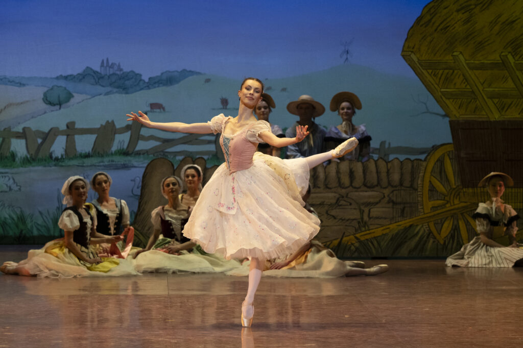 Bleuenn Battistoni balances in back attitude, arms open in offering to the audience. She wears a pale pink dress that falls just below the knee over pink tights and pointe shoes. A pastoral scene is visible in the background, a handful of dancers sitting or standing as they watch her perform.