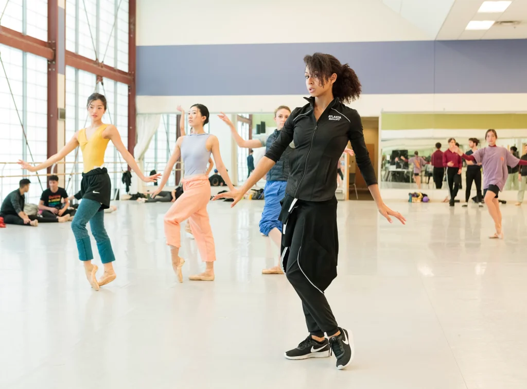 Claudia Schreier demonstrates a pose with a beveled foot and flexed hands to pointe shoe wearing dancers behind her.