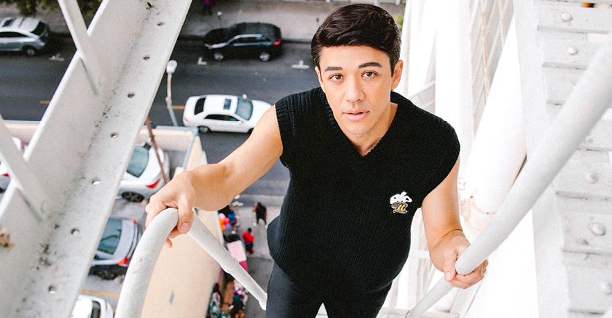 Kyle Hanagami looks curiously up at the camera as he climbs a white-painted fire escape, people and cars visible in the background far below.