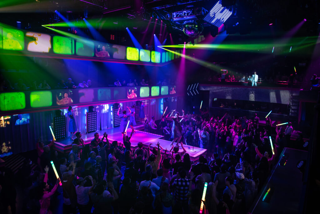 neon lights, glow sticks, and a large crowd dancing in a space with a catwalk down the center