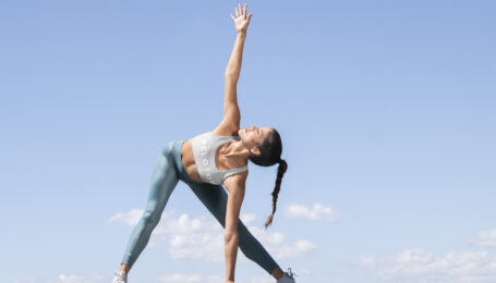 a woman stretching with the sky as her background, wearing blue leggings and a Peloton sports bra