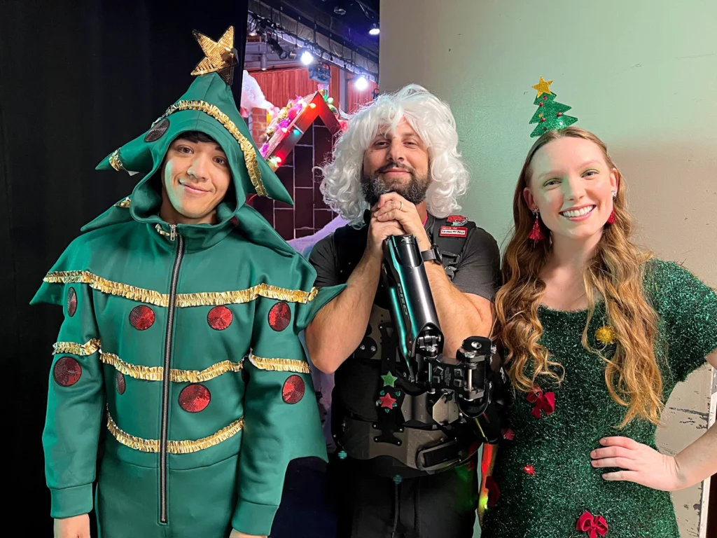 Kyle Hanagami, dressed in a Christmas tree onesie, gives a dopey smile to the camera. Beside him, a camera man wears a silly white wig and a young woman is dressed in a textured green dress adorned with little red bows.